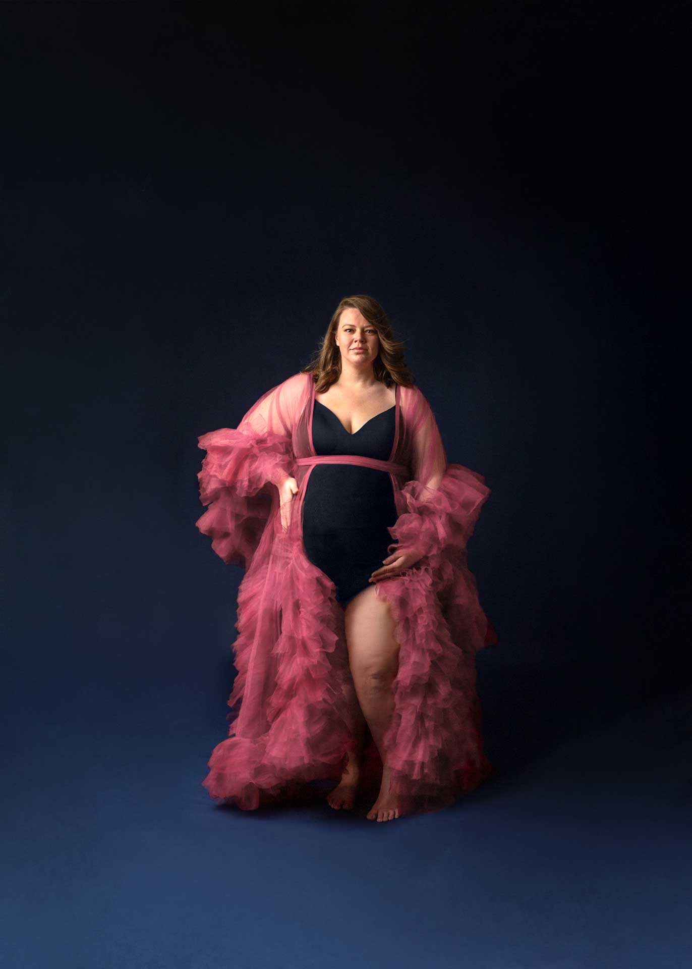 plus sized mom in pink tulle robe on dark blue backdrop minneapolis maternity photographer