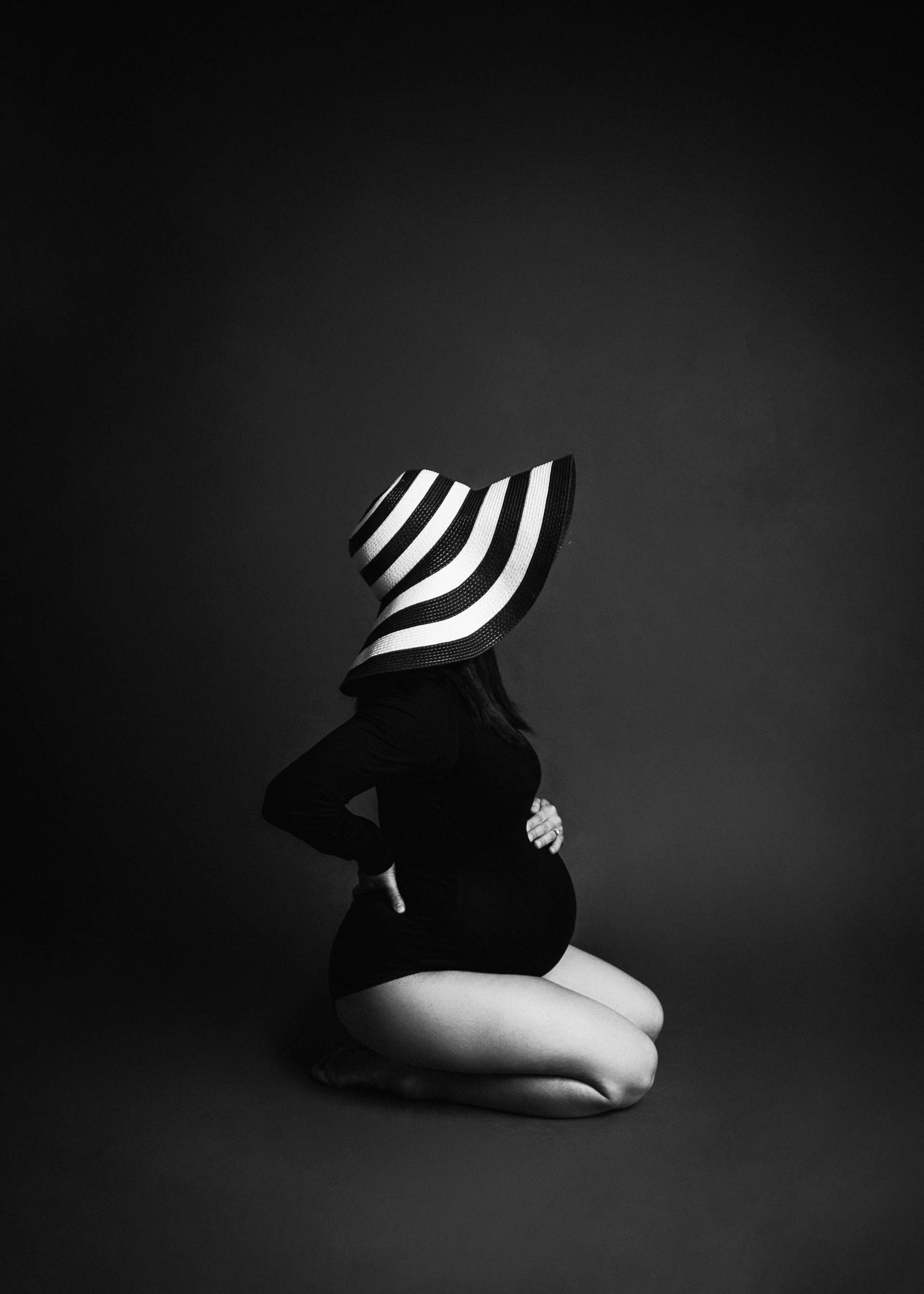 maternity in black body suit and black and white striped hat on gray backdrop maternity photography studio in minneapolis mn