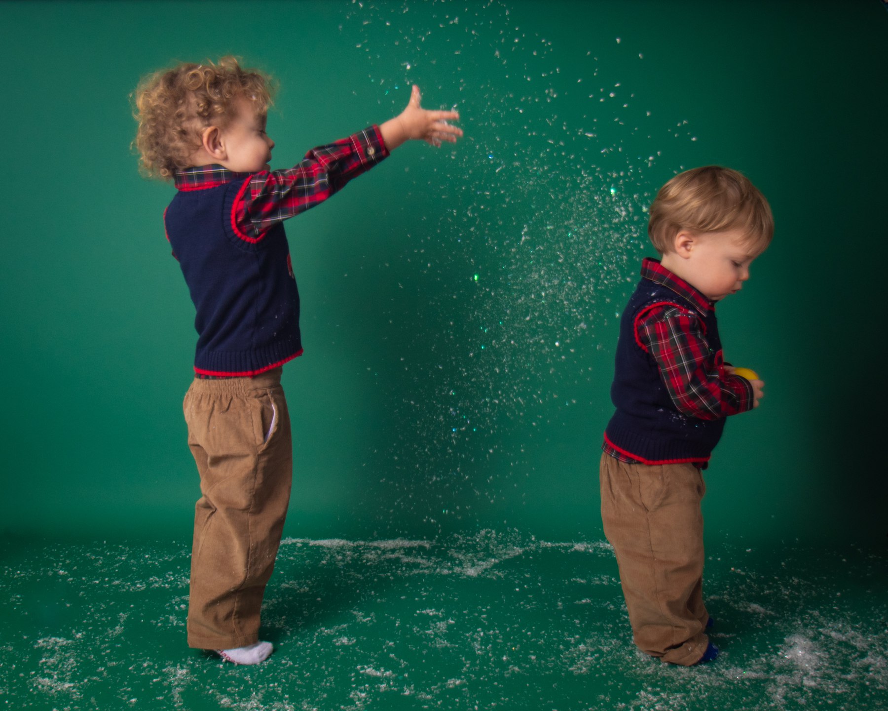 two toddler boys in christmas outfits tossing snow on green backdrop