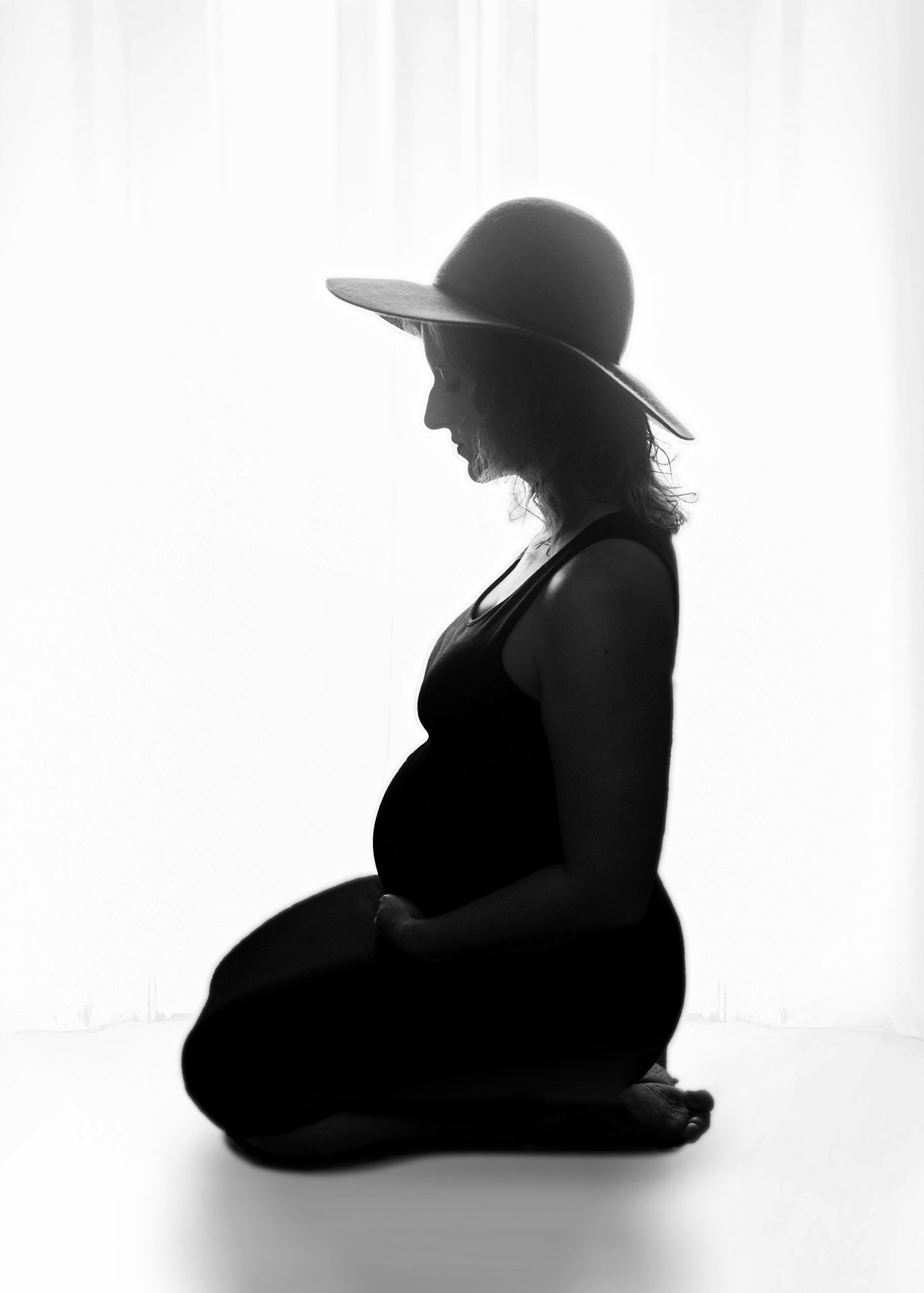maternity model in black dress and boho hat silhouetted against white curtains minneapolis maternity photography session